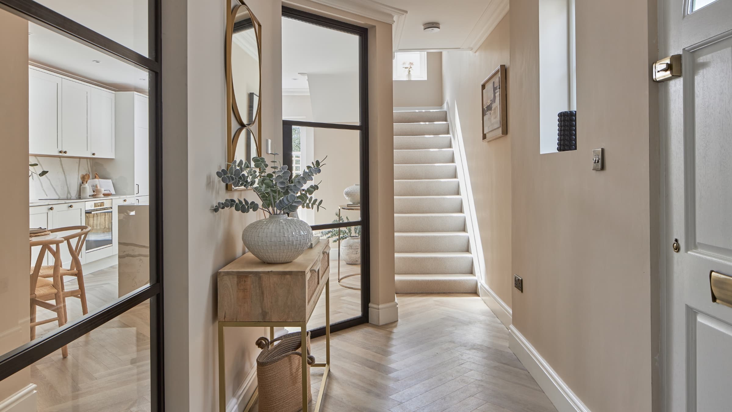 Professionally staged hallway leading to staircase. The hallway features glass partition walls looking into the kitchen area, mirrors for added light, designer furniture with decorative plant pots added. Light and airy feel.