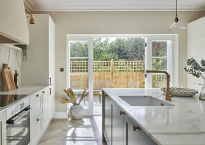 Staged home kitchen with open french doors looking into garden area with outdoor seating. Kitchen island in focus with decorative ornaments, minimalist style. Light and airy feel with gold accents. Plants carefully placed for added homely feel.
