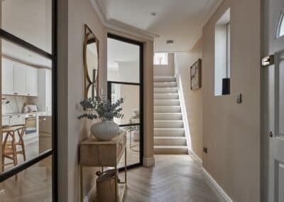Professionally staged hallway leading to staircase. The hallway features glass partition walls looking into the kitchen area, mirrors for added light, designer furniture with decorative plant pots added. Light and airy feel.